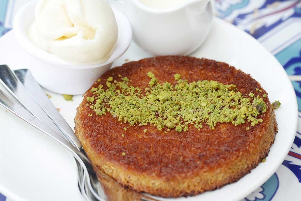 Kunafa with pistachios and a bowl of cream served on a white platter 