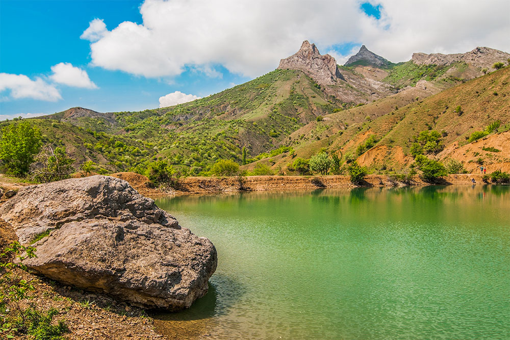 Mountain lake with bright green water, a large stone in the foreground on shore.