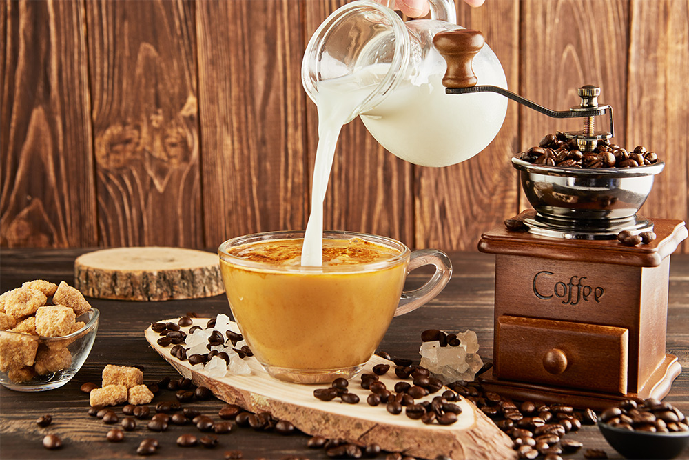 Pouring coffee from a glass jug of milk into a glass cup with coffee, a vintage coffee grinder, and lump sugar on a wooden background.