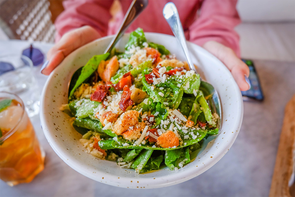 Close up and selective focus of hands holding Caesar salad in white bowl with fork and spoon in a restaurant or dining room in healthy food concept.