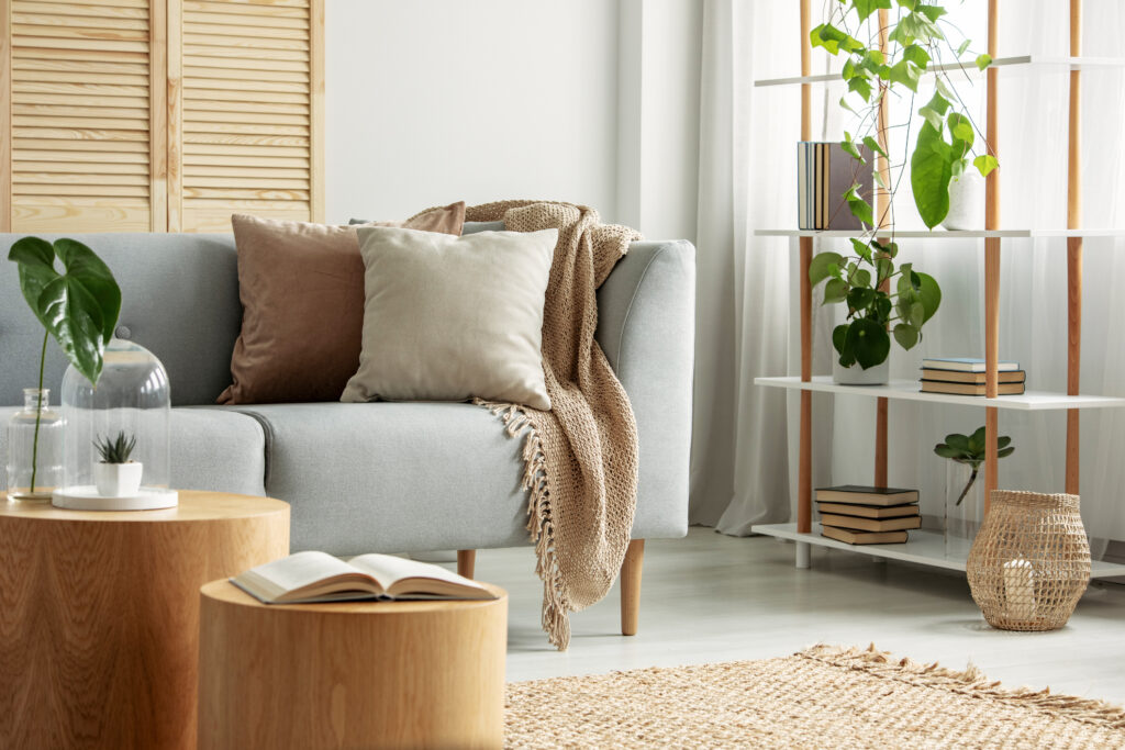 A cozy, well-staged living room with a light grey sofa adorned with neutral-colored pillows and a soft throw blanket. A small wooden coffee table holds decorative plants and an open book. In the background, a bookshelf with plants and neatly arranged books adds to the welcoming ambiance. This image highlights how effective home staging can create an inviting atmosphere, appealing to potential buyers during a real estate sale.