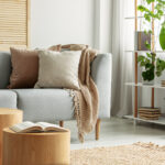 A cozy, well-staged living room with a light grey sofa adorned with neutral-colored pillows and a soft throw blanket. A small wooden coffee table holds decorative plants and an open book. In the background, a bookshelf with plants and neatly arranged books adds to the welcoming ambiance. This image highlights how effective home staging can create an inviting atmosphere, appealing to potential buyers during a real estate sale.