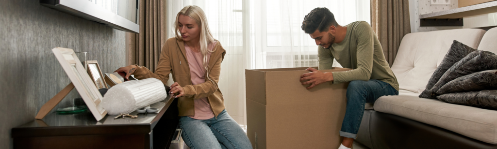 A couple unpacking boxes in their new home, carefully arranging personal items while settling in. This image could be used in a blog post for Property Finder’s Buyers and Renters guides, illustrating the moving process and making a new house feel like home.