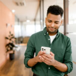 A real estate agent in a green shirt smiles while using his smartphone, standing in a modern office. This image illustrates the convenience of centralizing client communications on platforms like WhatsApp, enabling real estate agents to efficiently manage contacts and streamline interactions.