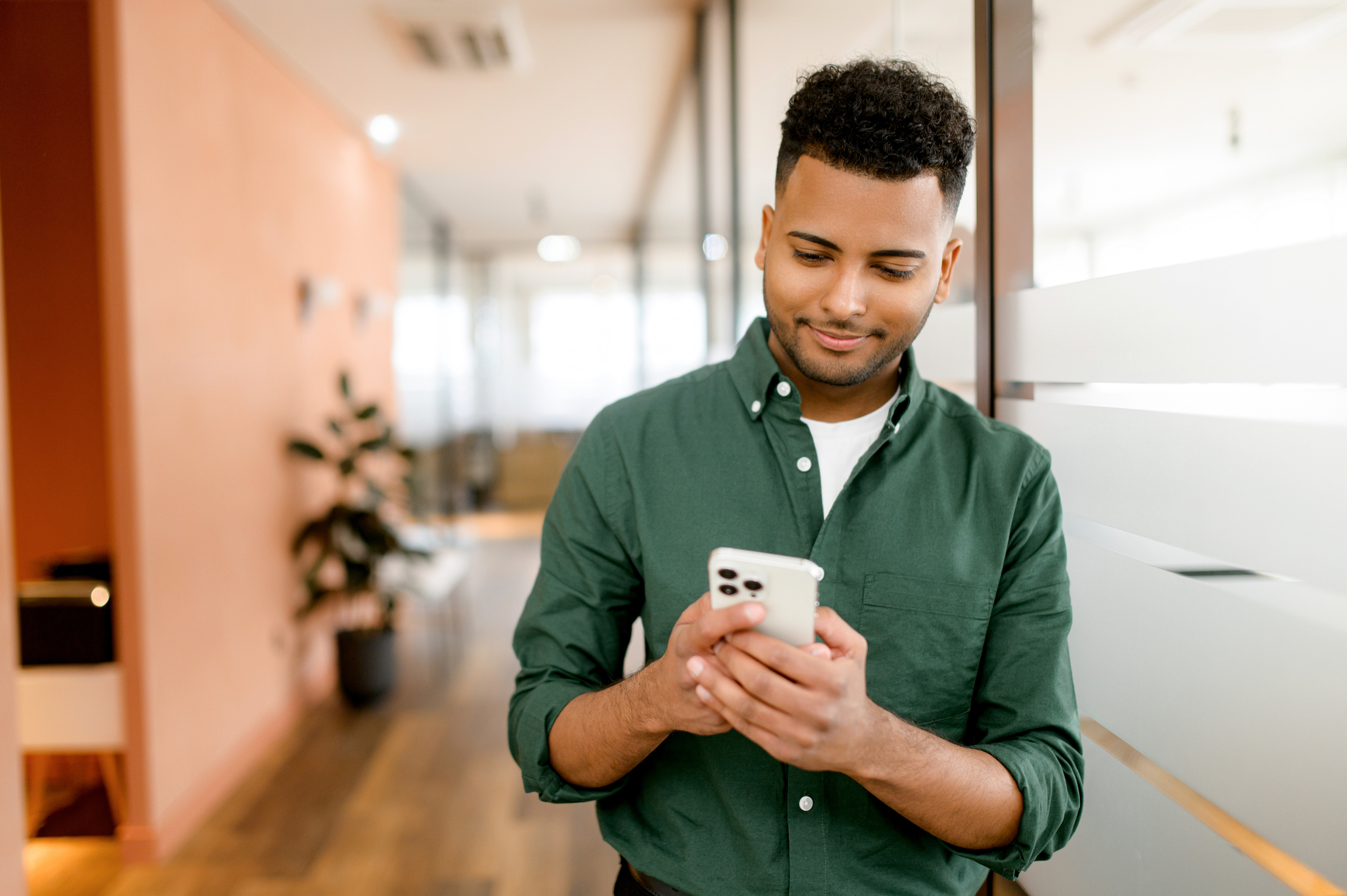 A real estate agent in a green shirt smiles while using his smartphone, standing in a modern office. This image illustrates the convenience of centralizing client communications on platforms like WhatsApp, enabling real estate agents to efficiently manage contacts and streamline interactions.