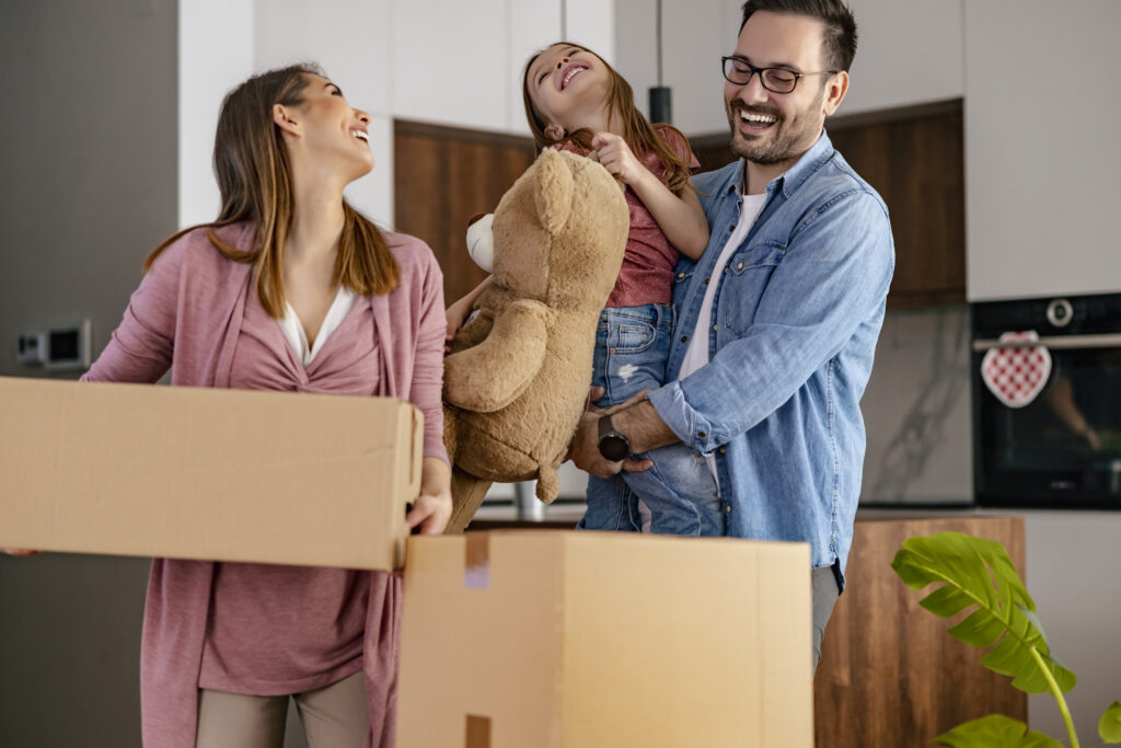 A smiling family moving into a new home, with the mother carrying a box, the father holding their child and a stuffed toy, reflecting the excitement and joy of moving to a new house or apartment in the UAE.