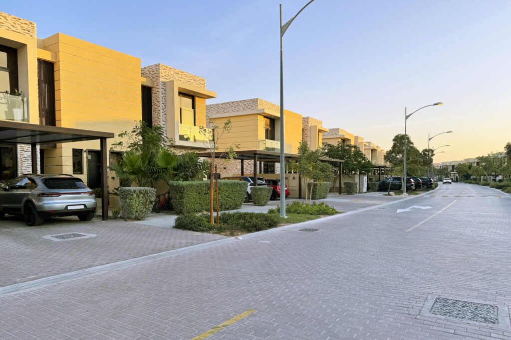 A beautiful residential street featuring modern townhouses in Dubai. The homes have contemporary architecture with flat roofs, large windows, and driveways. Several cars are parked outside, and the area is landscaped with lush greenery, well-maintained hedges, and trees, all under a clear summer sky.