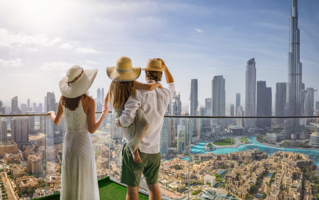 A group of three people, dressed in summer clothing and wide-brimmed hats, stand on a balcony overlooking the modern skyline of Dubai. The iconic Burj Khalifa and other tall skyscrapers rise in the distance, with a clear blue sky and the city's urban landscape below them. The scene reflects a luxurious and relaxed lifestyle in Dubai.