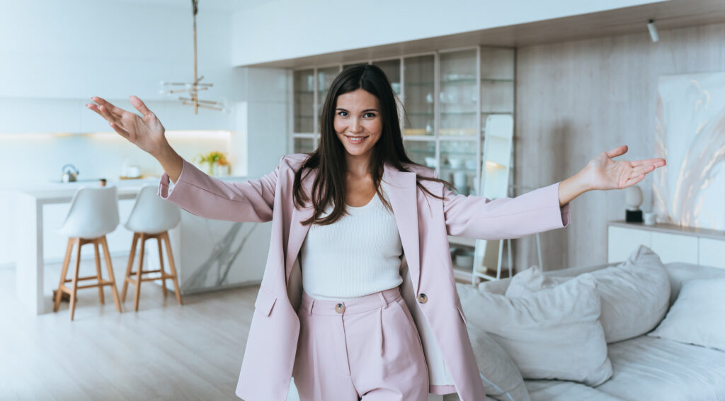 Cheerful real estate agent woman in a suit spreads her arms and welcoming people to the open house in a new real estate development.